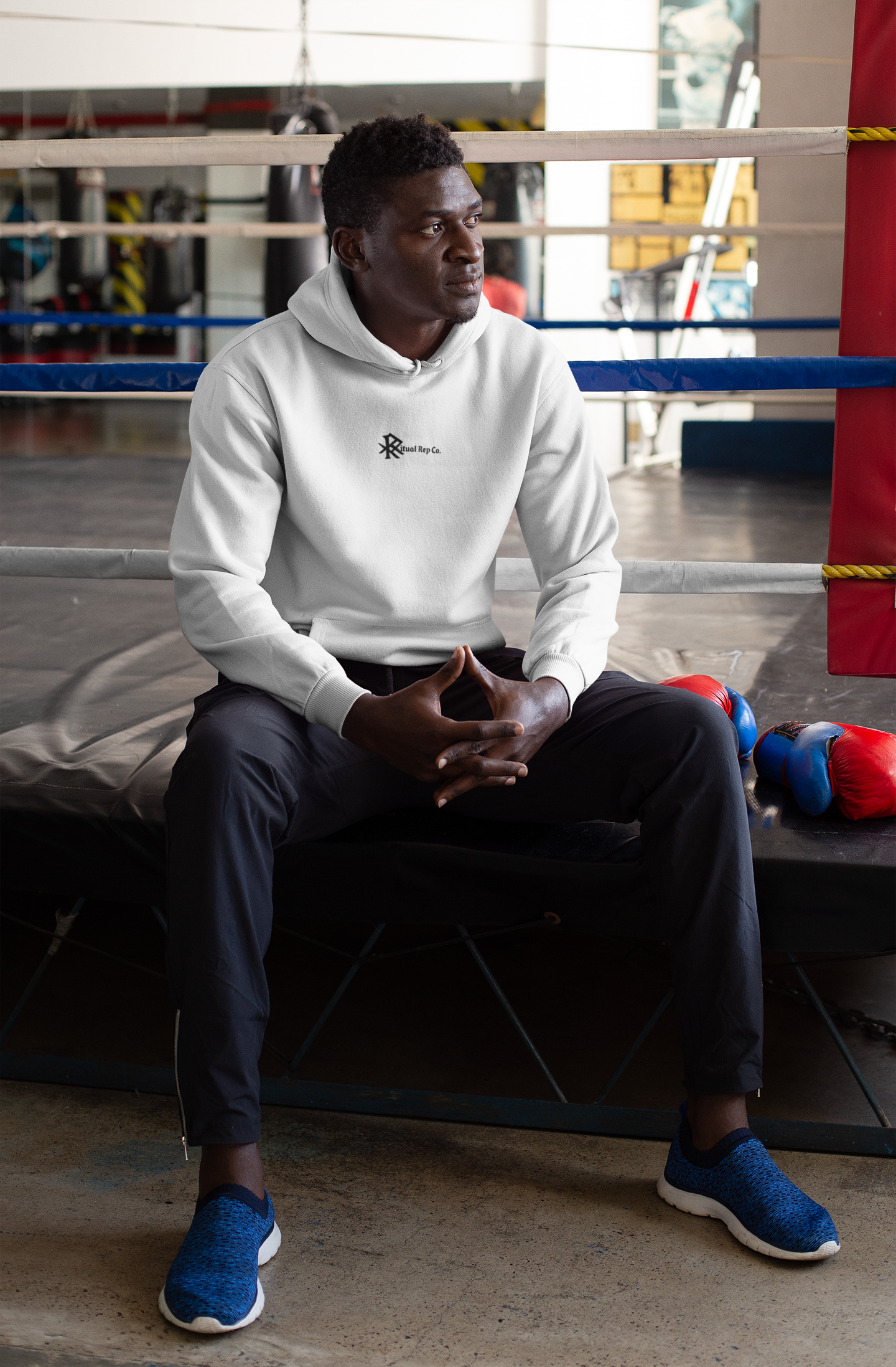Model seated in gym wearing white Initiate Hoodie—Ritual emblem visible on front.